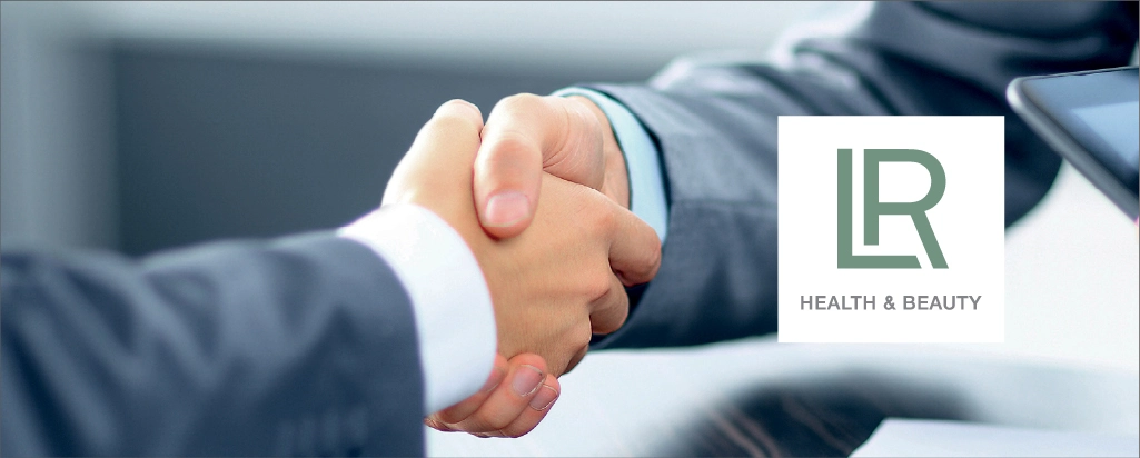 Poignée de main entre deux personnes en costume avec le logo de LR Health & Beauty en arrière-plan, symbolisant l'accompagnement approfondi et le soutien personnalisé des clients.
